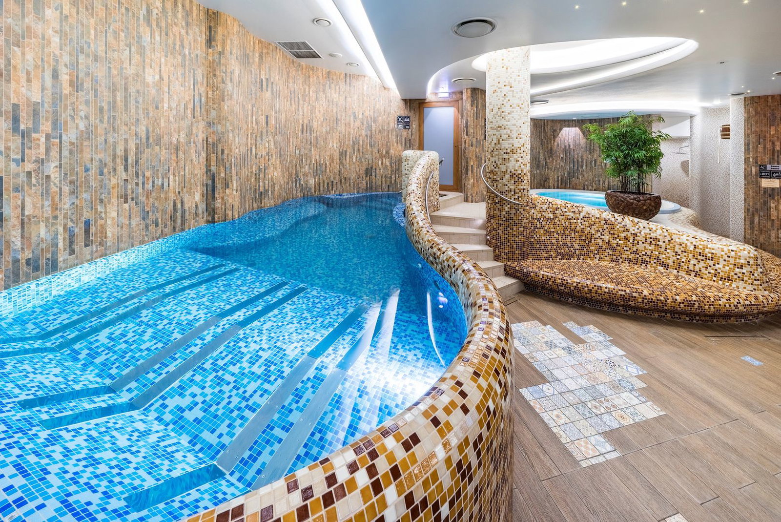 Indoor luxury spa pool with mosaic-tiled curved edge, submerged steps, whirlpool area and potted plant in a wellness centre