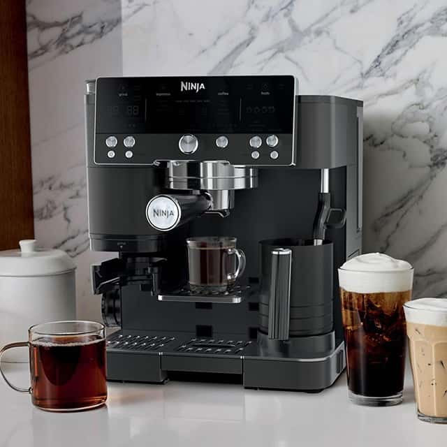 A sleek, black Ninja coffee machine sits on a kitchen counter with a marble backsplash. In front, there's a mug of black coffee and two tall glasses filled with coffee topped with foam. A white sugar bowl and a milk frother are also visible.