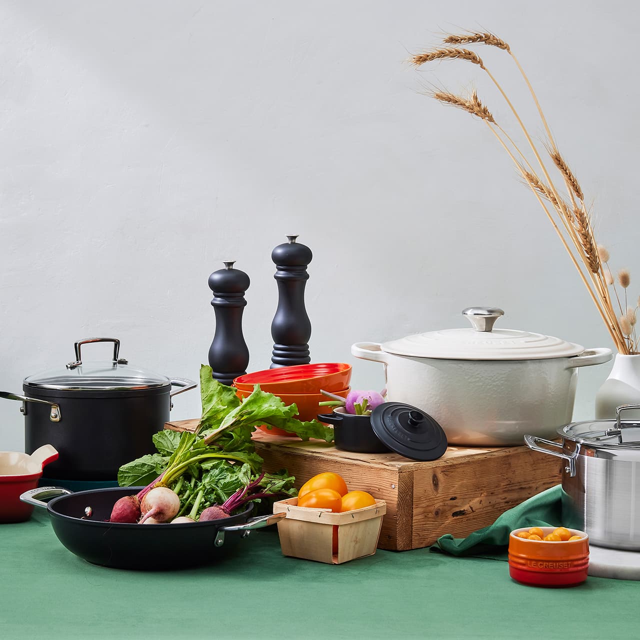 A kitchen scene featuring a variety of cookware, including a white casserole dish, a silver pot, and a black pan. Fresh vegetables like leafy greens and onions are on a wooden crate, along with oranges in a small box. Two black pepper mills and a bundle of dried wheat stalks complete the arrangement