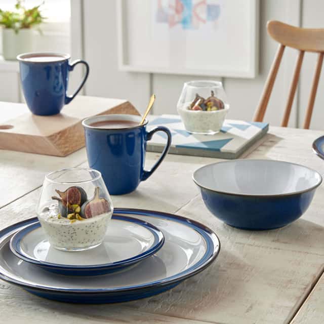 A dining table is set with blue and white crockery, including plates, bowls, and mugs. Two small glasses contain chia pudding topped with figs. The table is in a bright, airy room with a chair and framed pictures on the wall.