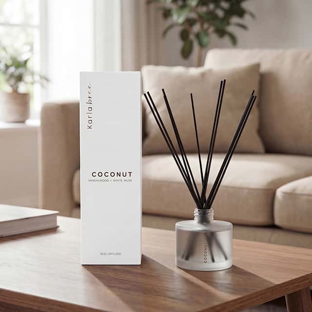 Reed diffuser with black sticks in a grey bottle labelled 'Coconut' sits on a wooden table next to its packaging. The box reads 'Coconut, Sandalwood, White Musk.' In the background, there's a beige sofa and a softly lit room with plants and large windows.