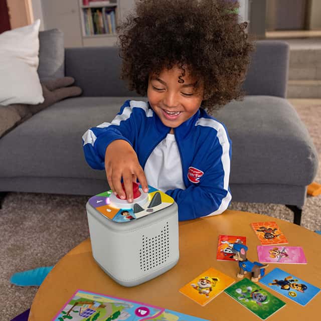 A young child with curly hair is smiling while placing a colourful figure on a cube-shaped audio device. The child is wearing a blue and white tracksuit. Various illustrated cards are spread out on the table, featuring cartoon animals. The setting includes a sofa and soft furnishings in a cosy room.