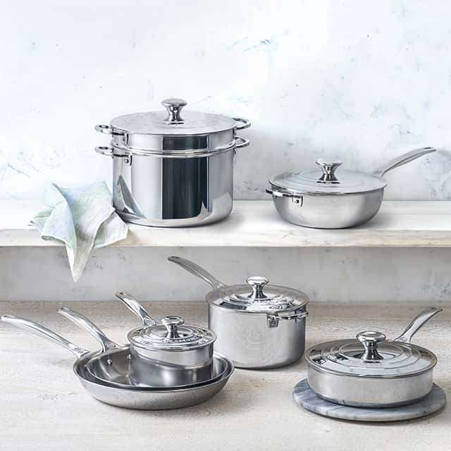 A collection of shiny stainless steel cookware is arranged on two wooden shelves against a light-coloured wall. The set includes saucepans, a stockpot, and frying pans with long handles. A kitchen towel is casually placed beside the stockpot on the top shelf.