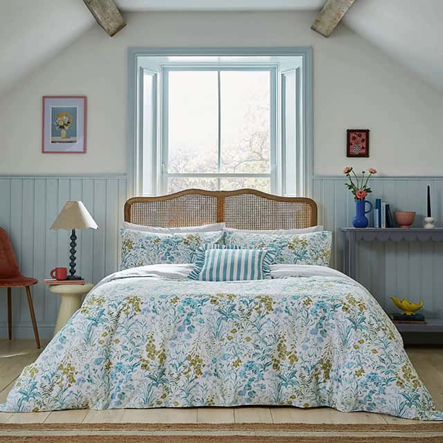 Attic bedroom with a large double bed featuring a floral duvet and pillows. The room has a sloped ceiling with wooden beams, a window with light blue shutters, and light grey panelled walls. A small wooden table with a lamp is on one side, and a shelf with vases and books is on the other. Warm light