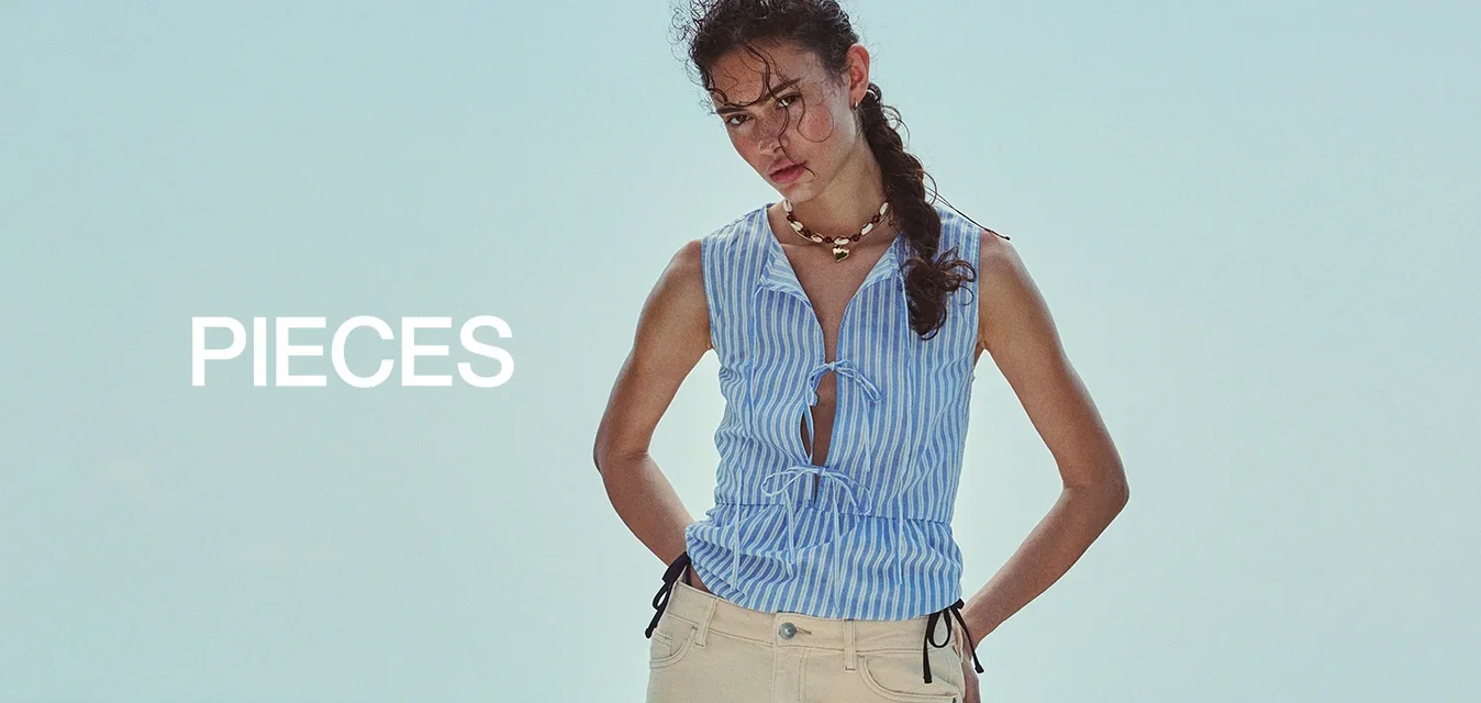 Vrouw met gevlochten haar staat op een houten dek bij de zee, draagt een mouwloos blauw gestreept topje, beige knielange shorts en zwarte slippers.