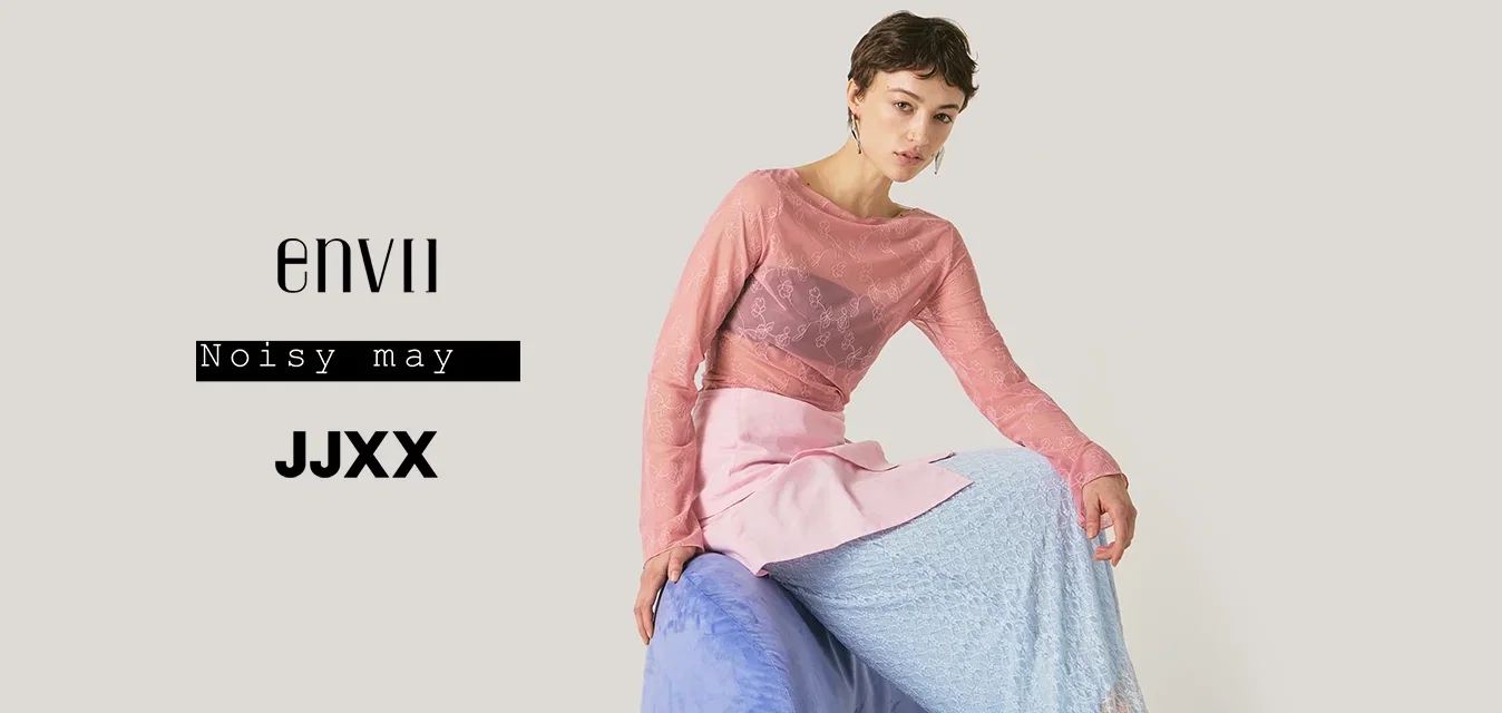 Jonge vrouw in gelaagde doorschijnende roze top en blauwe rok zittend op een gebogen paarse stoel tegen een effen lichte achtergrond.