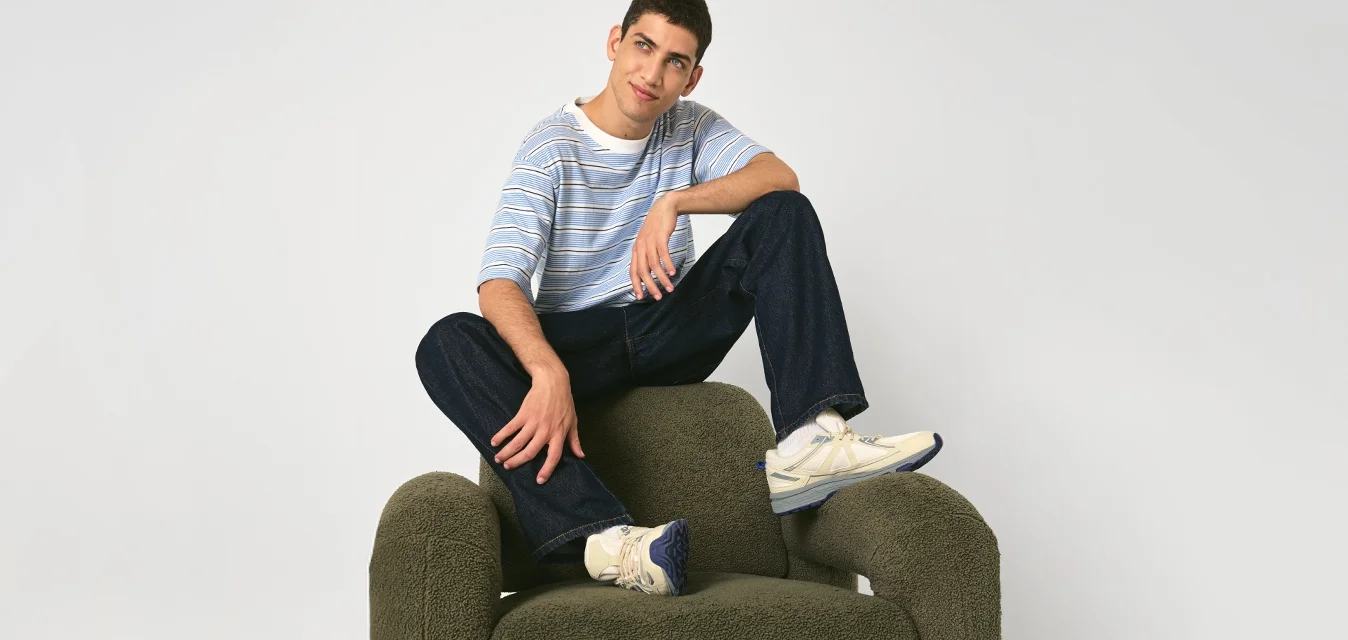 Jonge man in een gestreepte shirt en jeans die met gekruiste benen zit op een groene pluche fauteuil tegen een effen achtergrond.