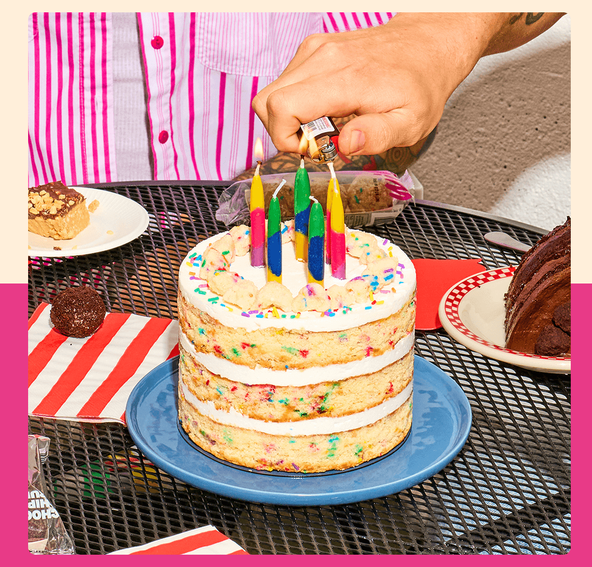A hand lights colorful candles on a sprinkle-filled Birthday Cake set on a blue plate, surrounded by assorted desserts on an outdoor table.