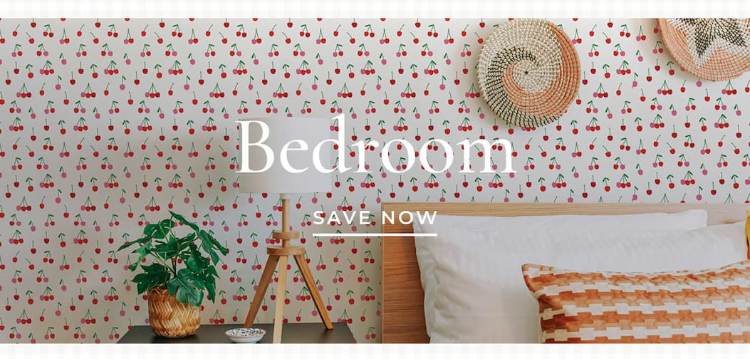 Image of Tempaper's Cherry Bomb wallpaper installed on a wall behind a bed and night stand with a lamp/ Text overlay. Bedroom Save Now.