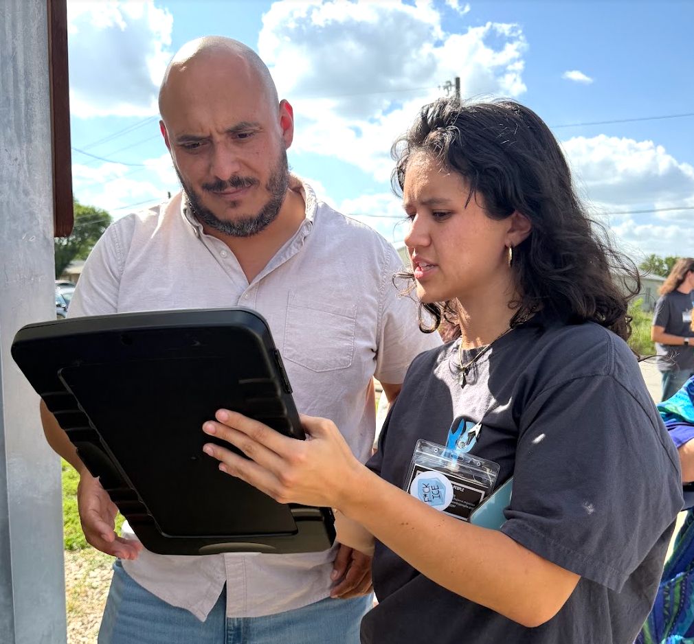 Two people looking at a clipboard that the person on the right is holding; they are standing outside