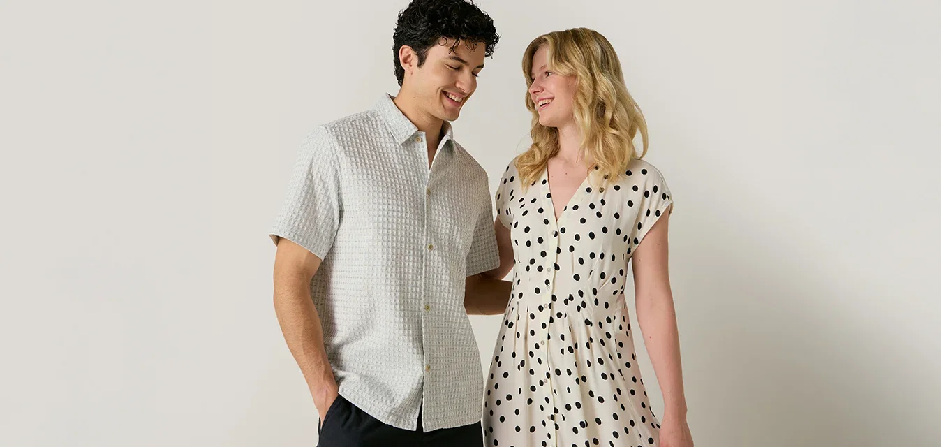 Un uomo in camicia a maniche corte e pantaloncini neri sta accanto a una donna con un vestito bianco con puntini neri, entrambi sorridono e si guardano.