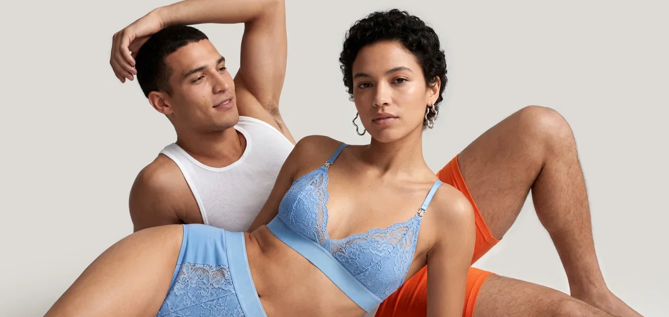 Un uomo con canottiera bianca e pantaloncini arancioni è sdraiato con una donna in lingerie azzurra, entrambi su uno sfondo chiaro e semplice.