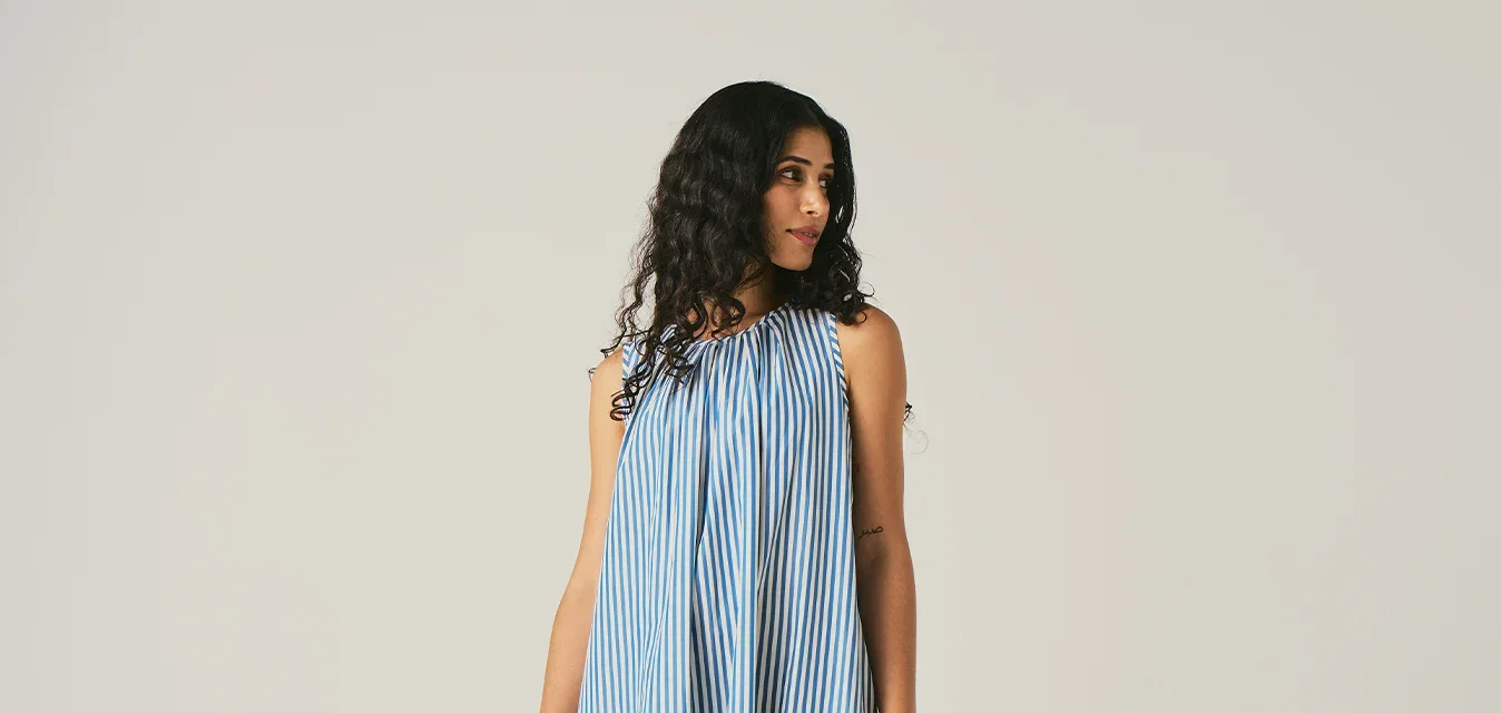 Femme aux longs cheveux bouclés portant une robe maxi sans manches à rayures verticales bleues et blanches, debout devant un fond clair uni.