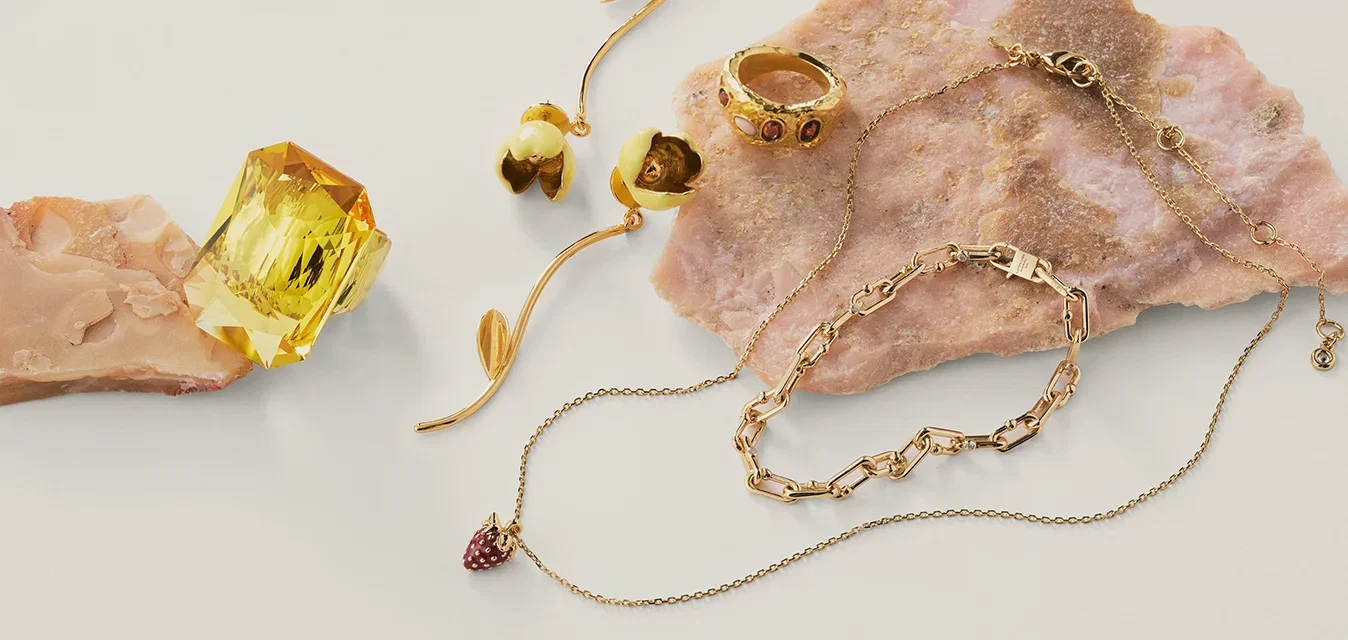 Bijoux en or variés comprenant une chaîne avec un petit pendentif, des bagues et des boucles d'oreilles disposés sur des pierres texturées rose pâle.