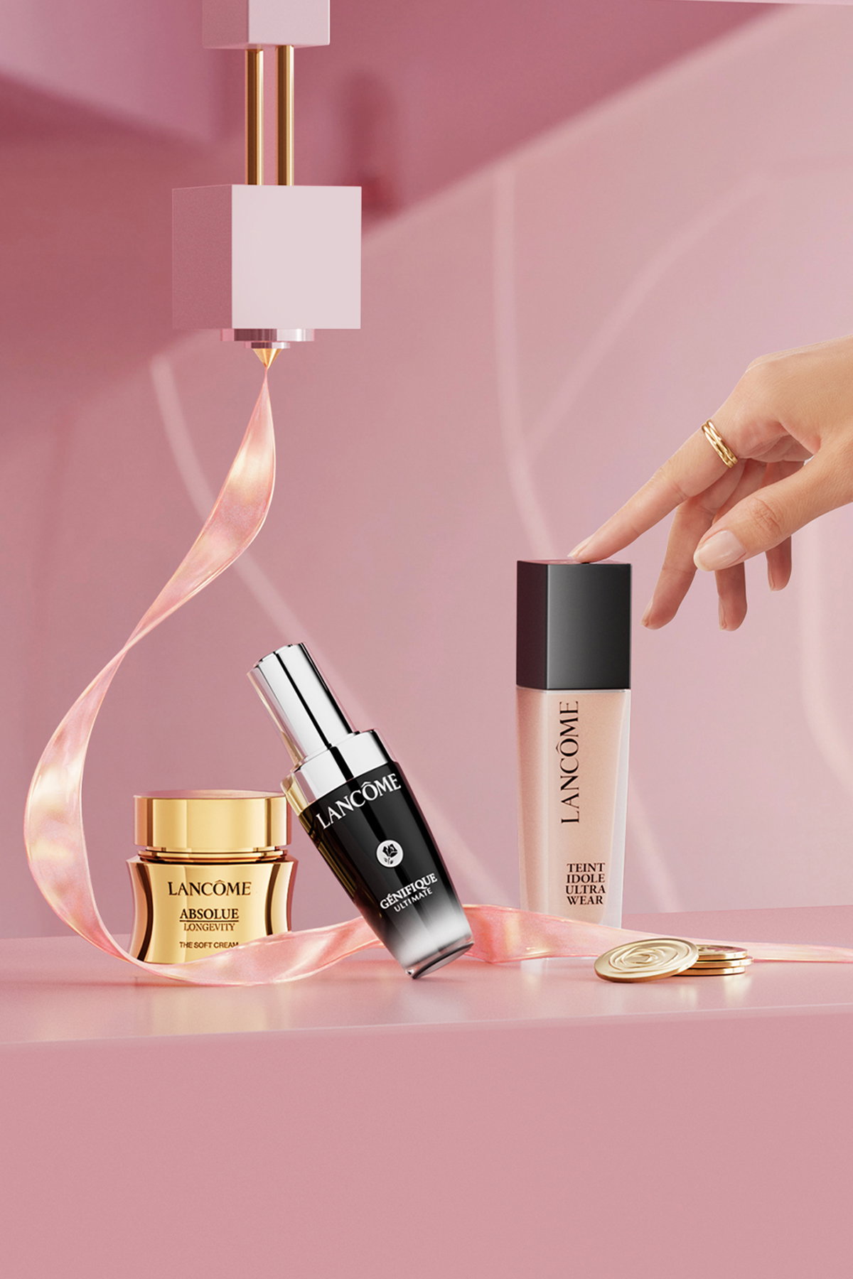Prodotti Lancôme su sfondo rosa con una mano che sfiora il flacone di fondotinta e un nastro decorativo.