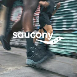 Zapatillas deportivas negras con un cuerpo texturizado y suelas de color gris claro en movimiento sobre el pavimento, con un fondo de graffiti urbano borroso.