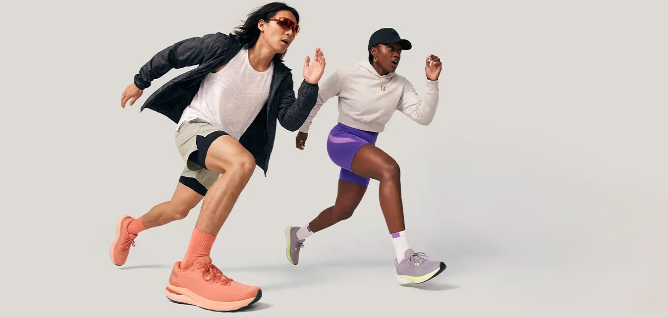 Dos corredores con ropa deportiva corren a toda velocidad hacia adelante sobre un fondo claro y liso, uno lleva zapatos naranjas y el otro pantalones cortos morados.
