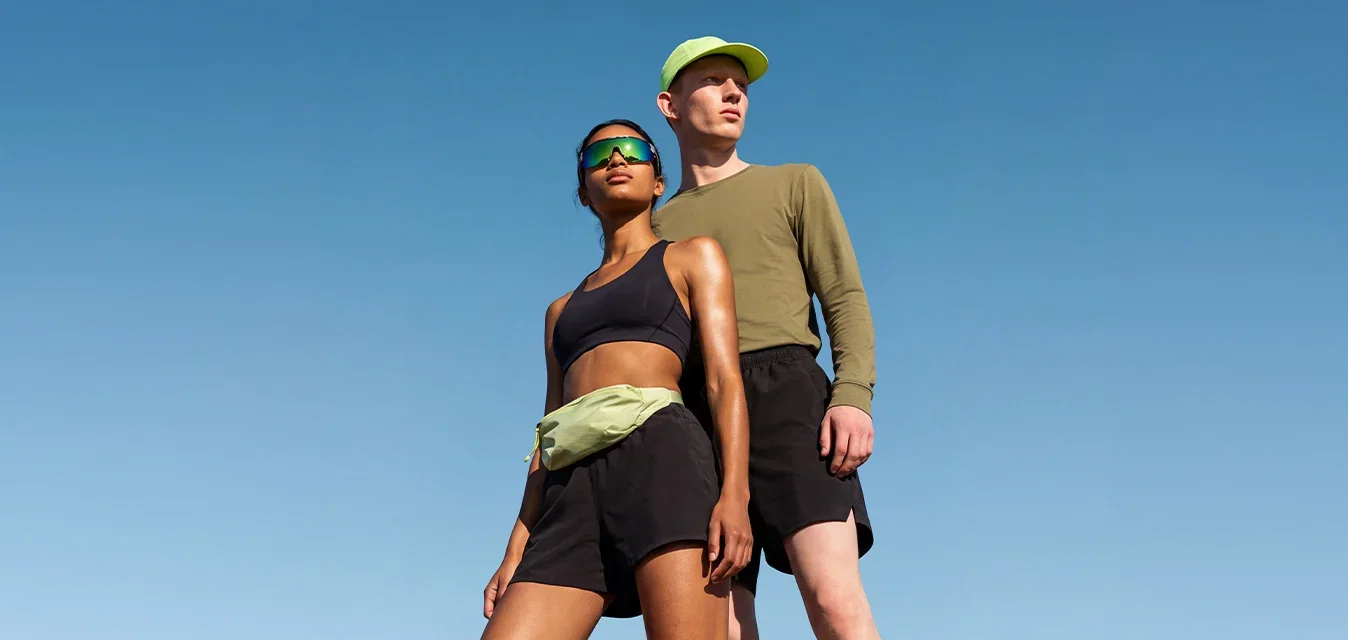 Dos excursionistas están de pie en la cima de una colina rocosa, vestidos con ropa deportiva, mirando a lo lejos bajo un cielo azul despejado.