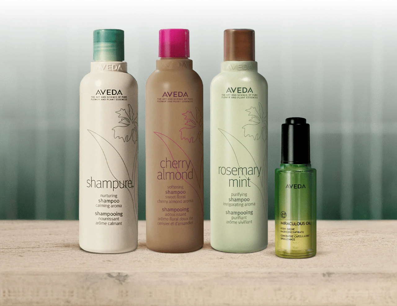 Three Aveda Shampoos lined up with a bottle of Miraculous Oil on a wooden surface, against an olive green background.
