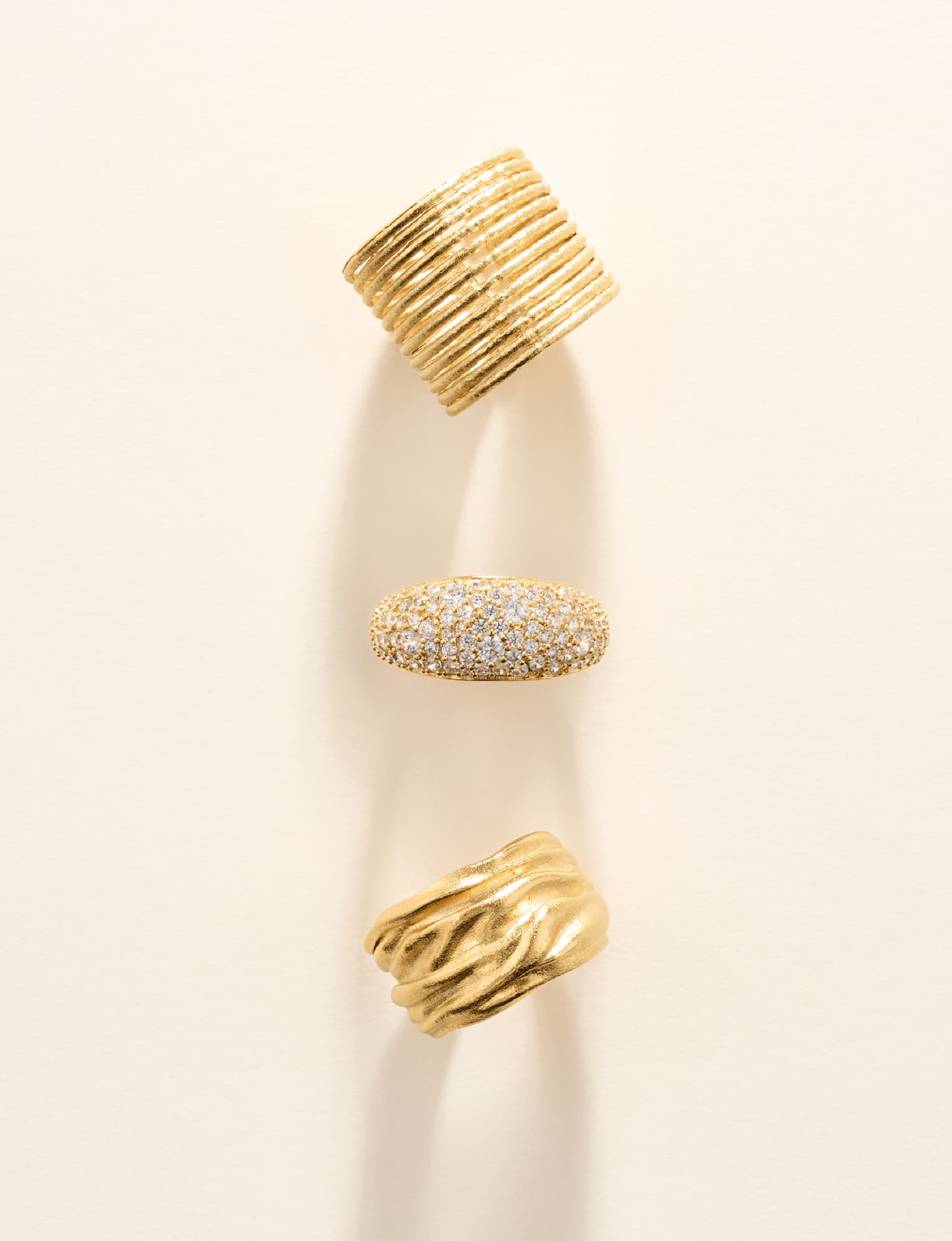 Three textured gold rings: a ribbed band, a crystal pavé dome and a draped wave band