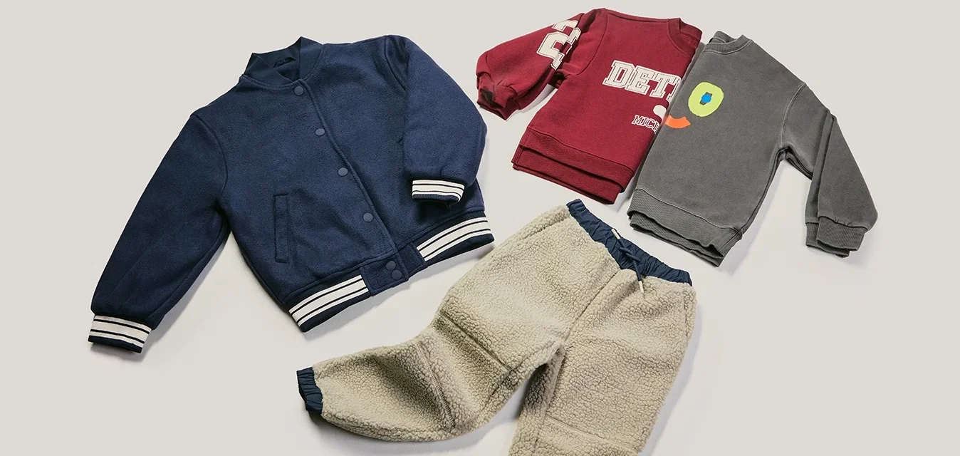 Marineblauwe varsityjas, rode sweatshirt met witte letters, grijze sweatshirt met kleurrijk gezicht en crèmewitte fleecebroek op een lichte achtergrond gerangschikt.
