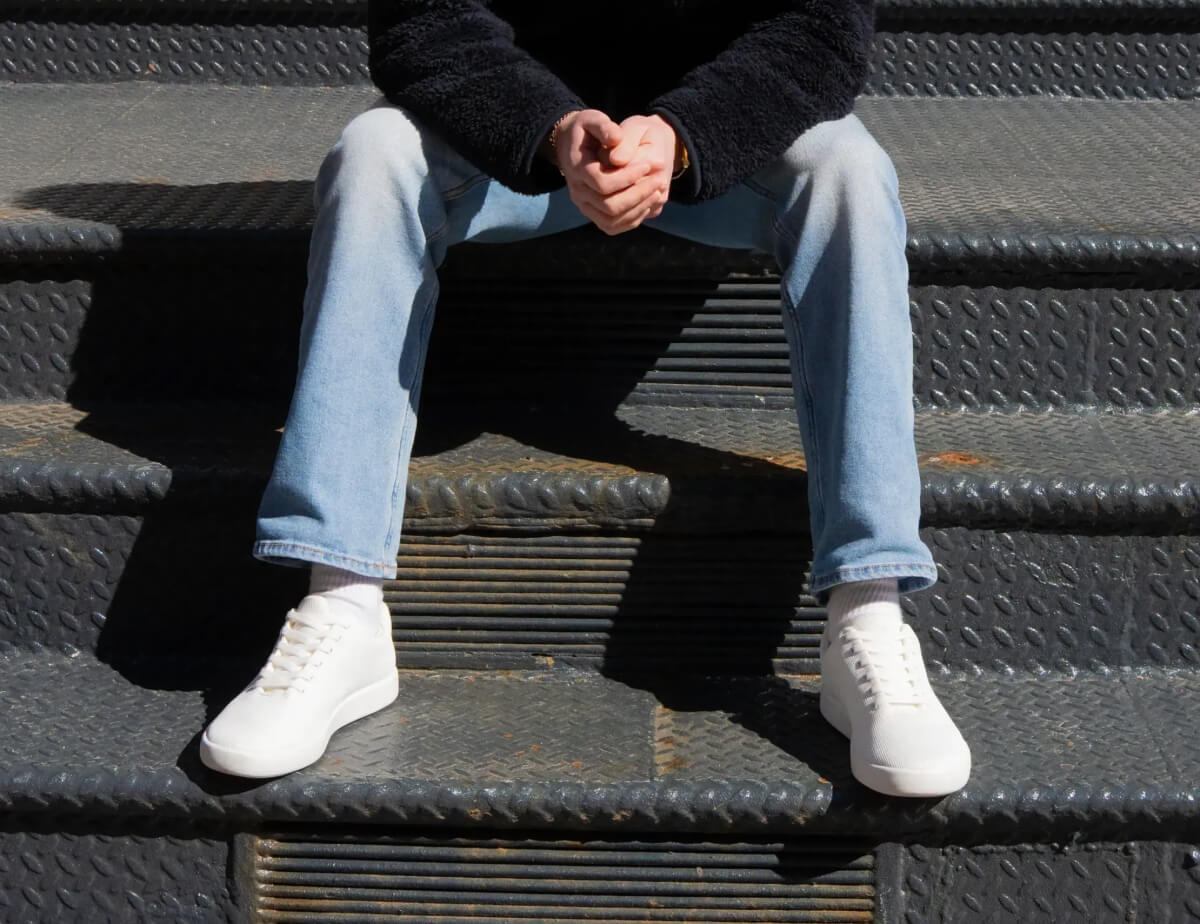 "Person in light jeans and white sneakers sits on metal stairs, hands clasped, casting a shadow in the sunlight."