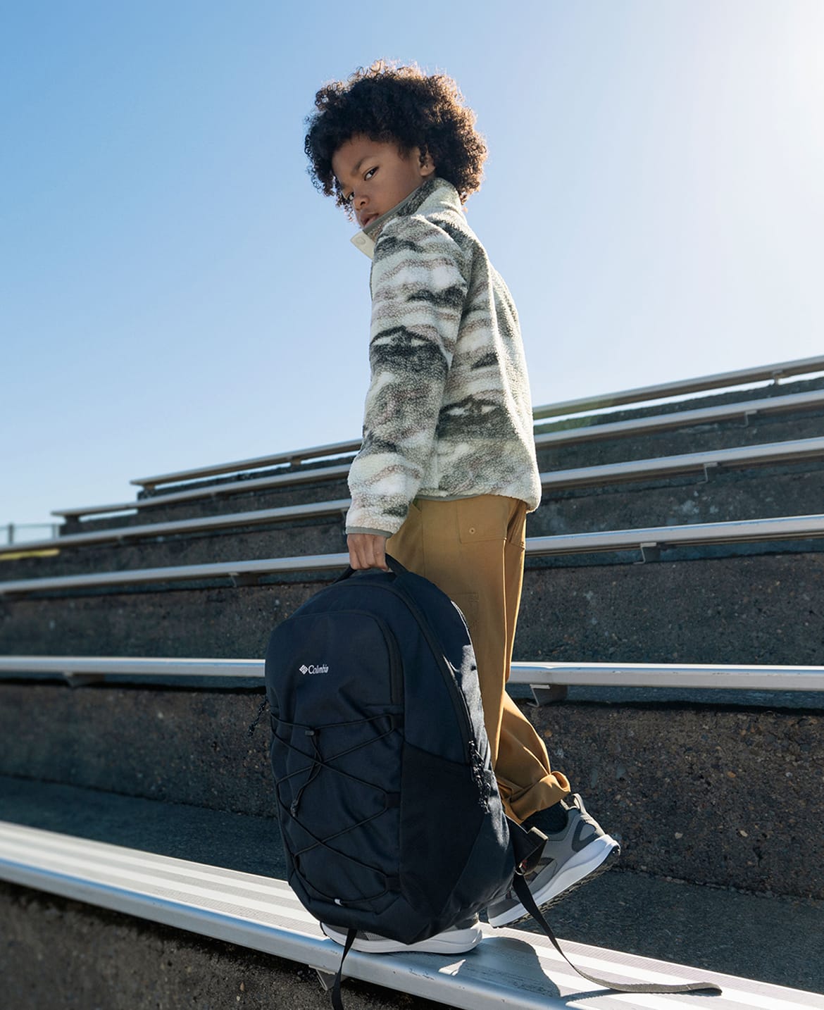 A little boy walking along bleachers, holding a black backpack.