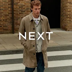Abrigo beige sobre un suéter gris y una camisa blanca, jeans oscuros, caminando por un paso de peatones. Fondo de pared de ladrillo, ambiente urbano casual.
