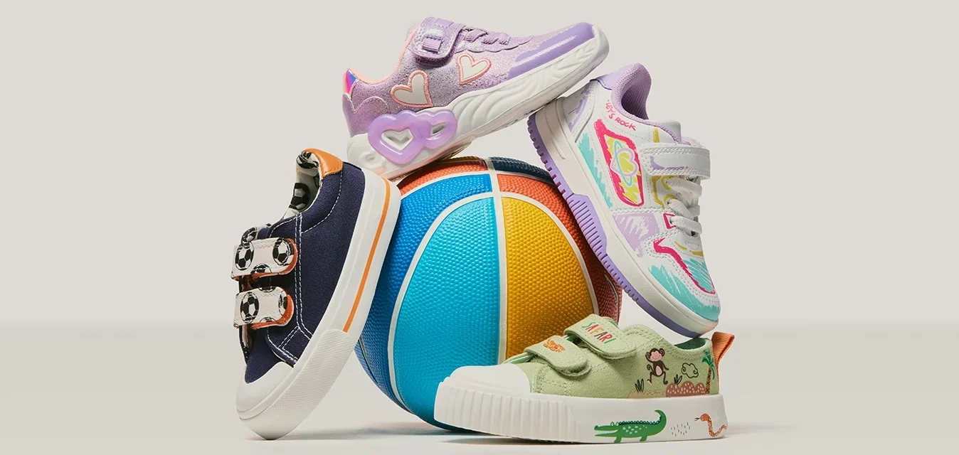 Cuatro zapatos infantiles de colores apilados alrededor de una pelota azul, amarilla y naranja sobre un fondo liso.