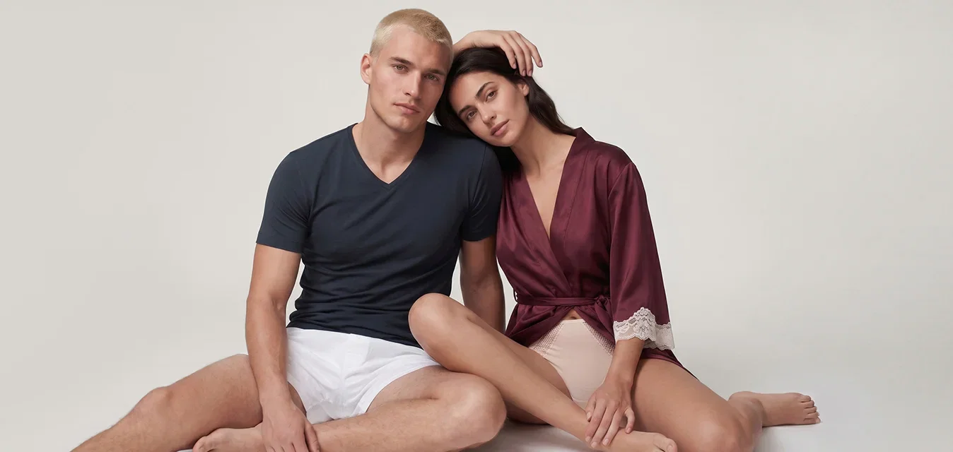 Jonge man en vrouw zitten op de vloer, de vrouw leunt met haar hoofd op de schouder van de man, beiden dragen casual minimalistische kleding.