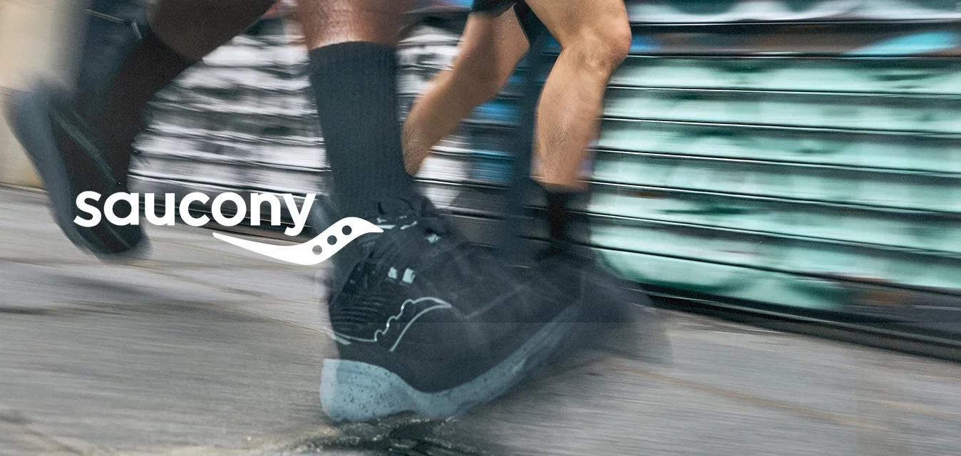 Zwarte sportieve schoenen met een gestructureerde body en lichtgrijze zolen, in beweging op het asfalt, met een vervaagde stedelijke graffiti-achtergrond.