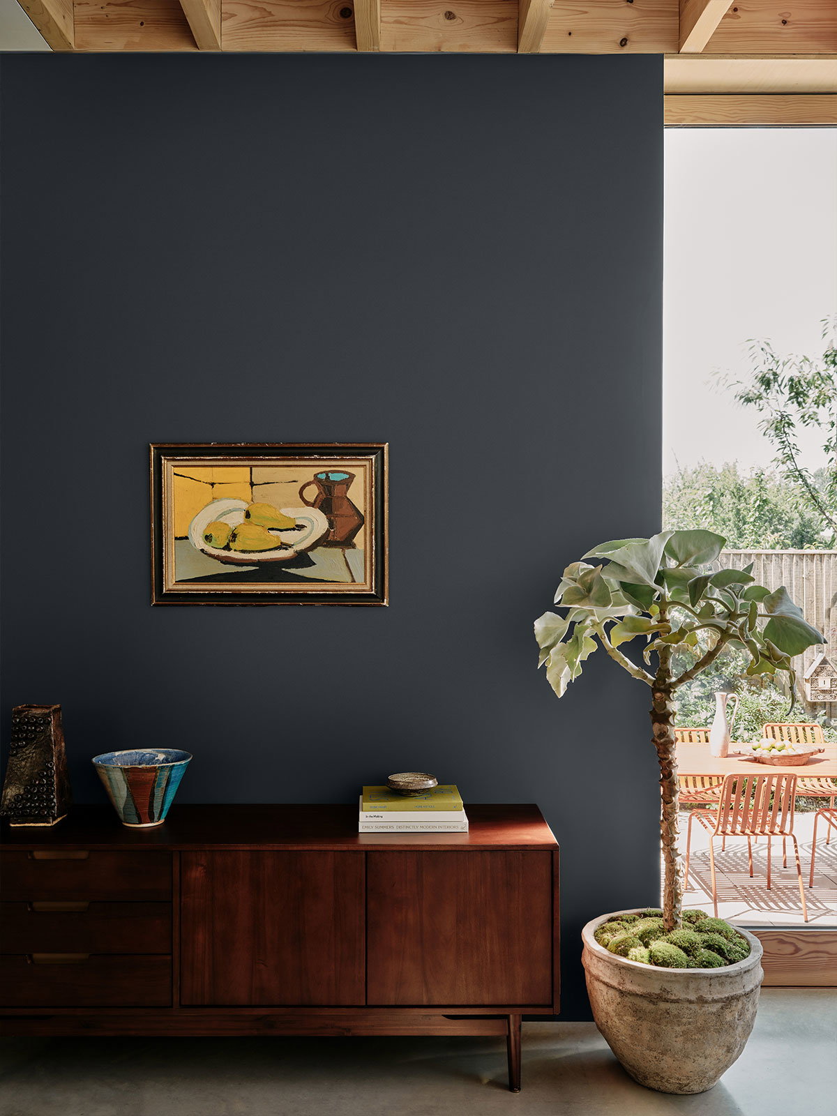 Mid-century style interior with dark blue wall, wooden sideboard, framed artwork, and potted plant.