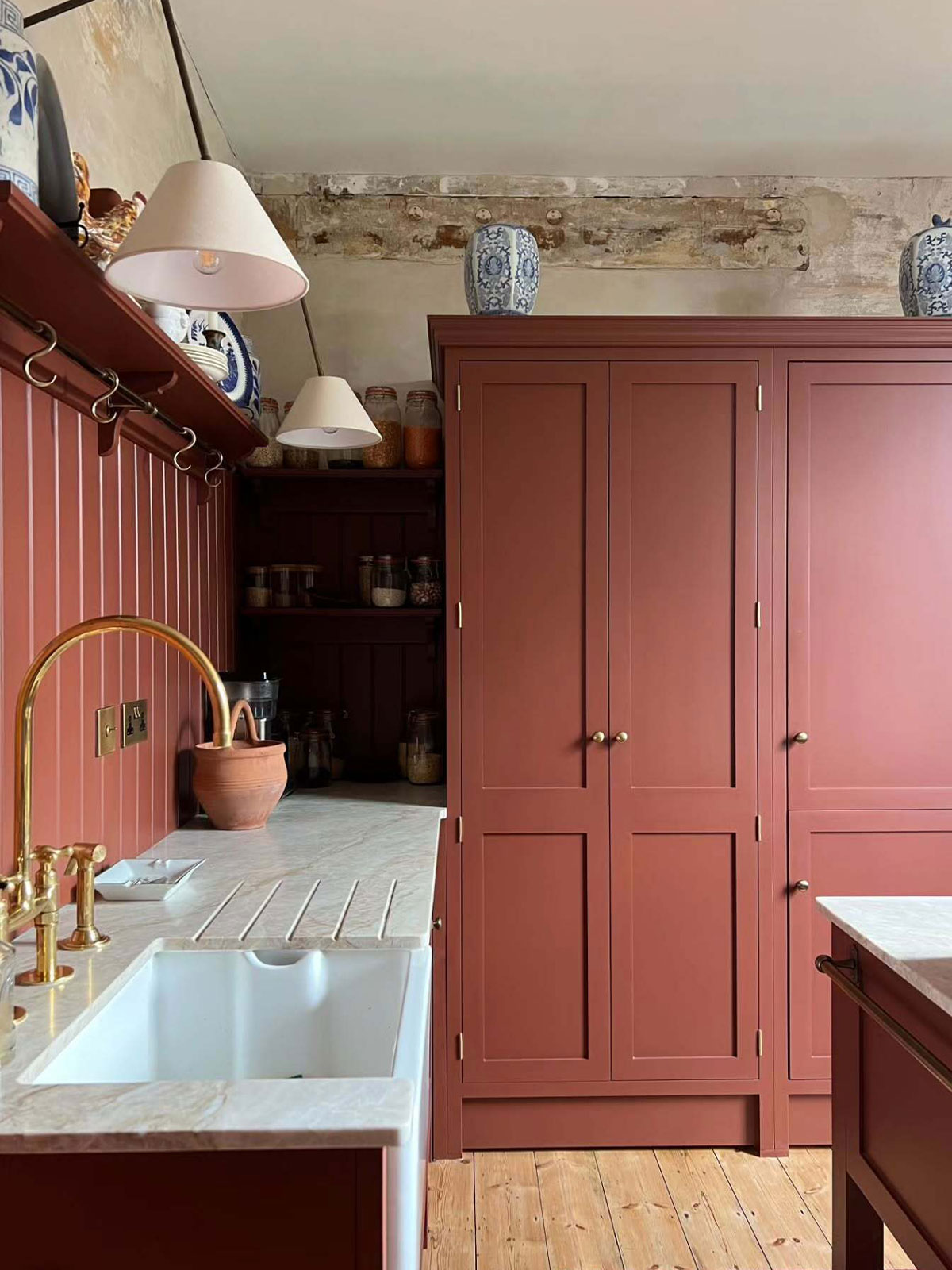 Warm kitchen with reddish-brown cabinets, marble worktops, brass tap, and blue patterned vases on top.