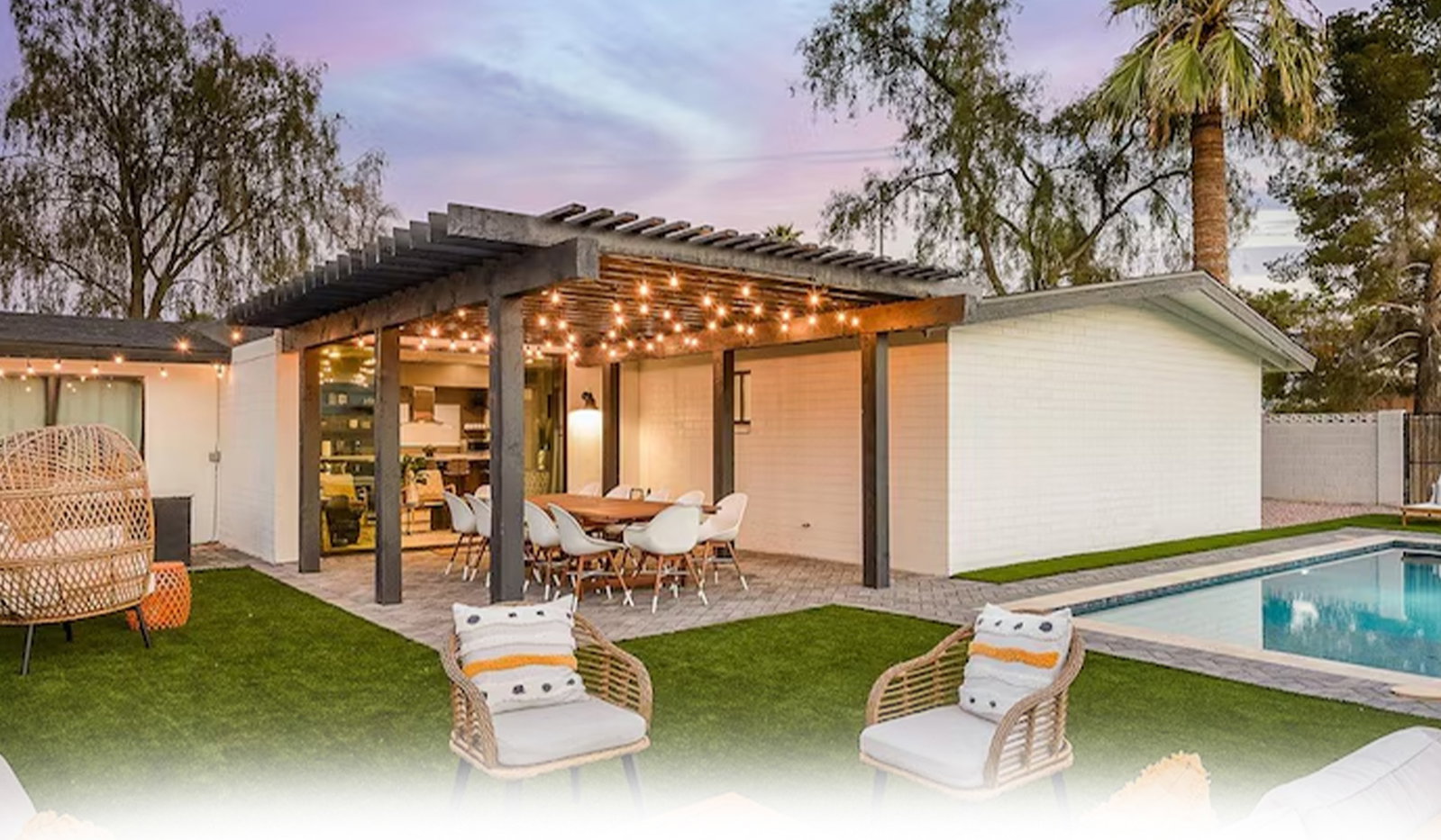 Modern backyard patio with outdoor dining area beneath a string-lit pergola and poolside lounge seating.