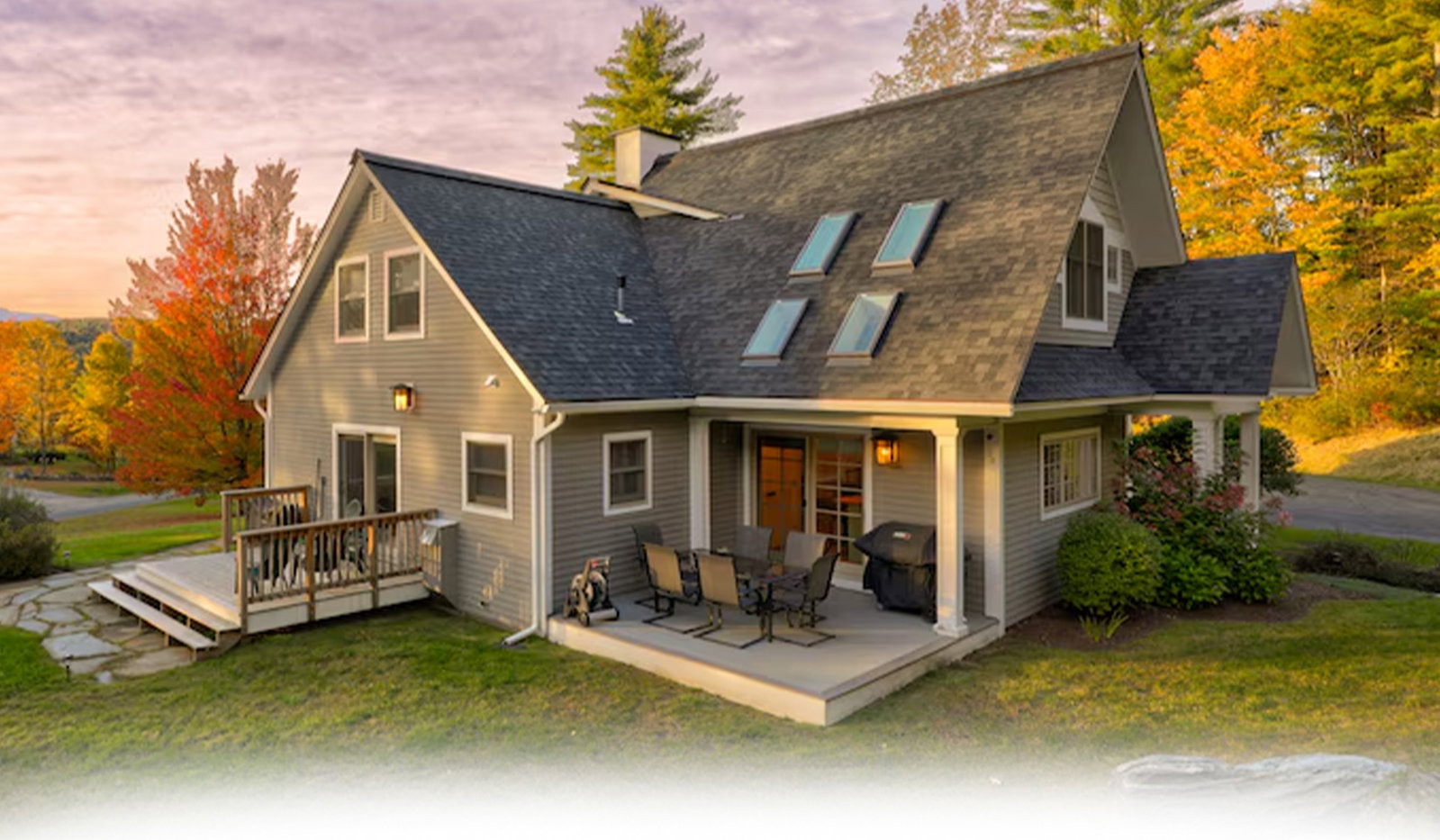 Gray contemporary farmhouse with skylights, porch, and autumn trees at sunset.