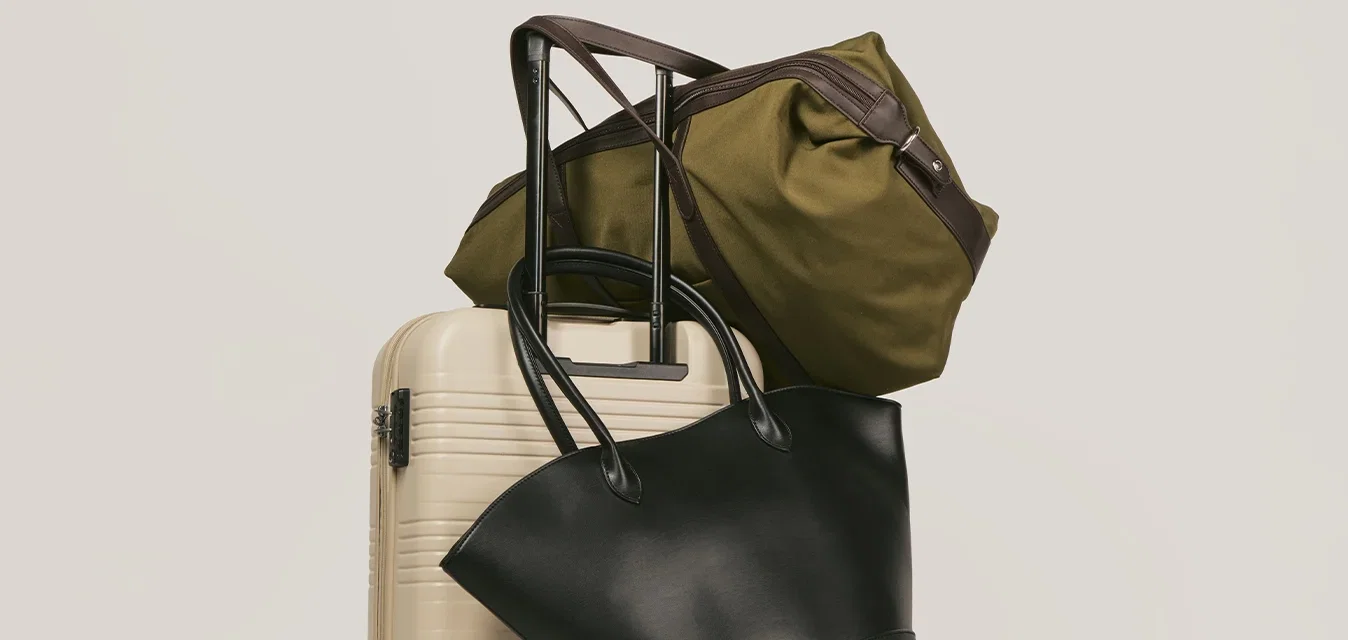 Valise à roulettes beige avec une poignée extensible portant un sac de sport olive et un sac à main en cuir noir sur un fond uni.