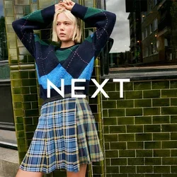 Pull en motif bleu, vert et marine avec une coupe ample, associé à une jupe plissée à carreaux avec des accents jaunes et une ceinture noire.