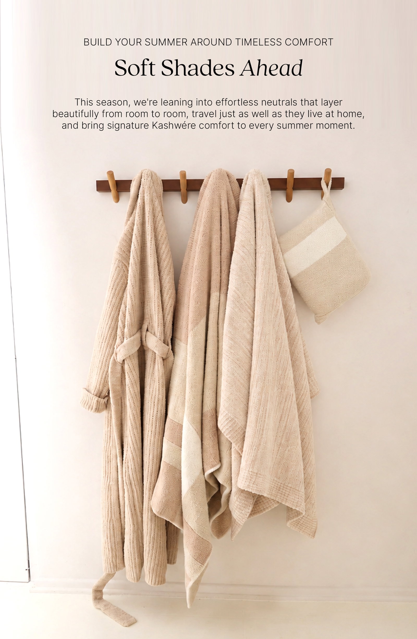 BUILD YOUR SUMMER AROUND TIMELESS COMFORT  Soft Shades Ahead  This season, we're leaning into effortless neutrals that layer beautifully from room to room, travel just as well as they live at home, and bring signature Kashwére comfort to every summer moment.  A wooden wall rack displays neutral Kashwére essentials including the Ojai Robe, Plaid Throw, Lightweight Striped Throw, and Stripe Travel Blanket in soft shades of cream, bone, and beige.