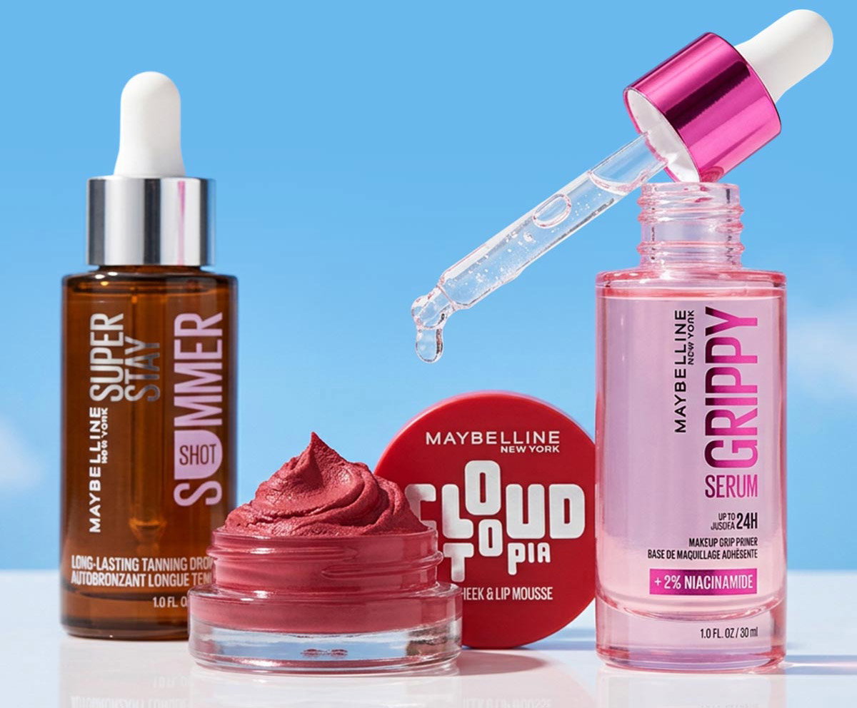 Maybelline New York product lineup against a blue sky: brown Super Stay Summer Shot tanning drops, red Cloudtopia Cheek & Lip Mousse, and pink Grippy Serum makeup primer.