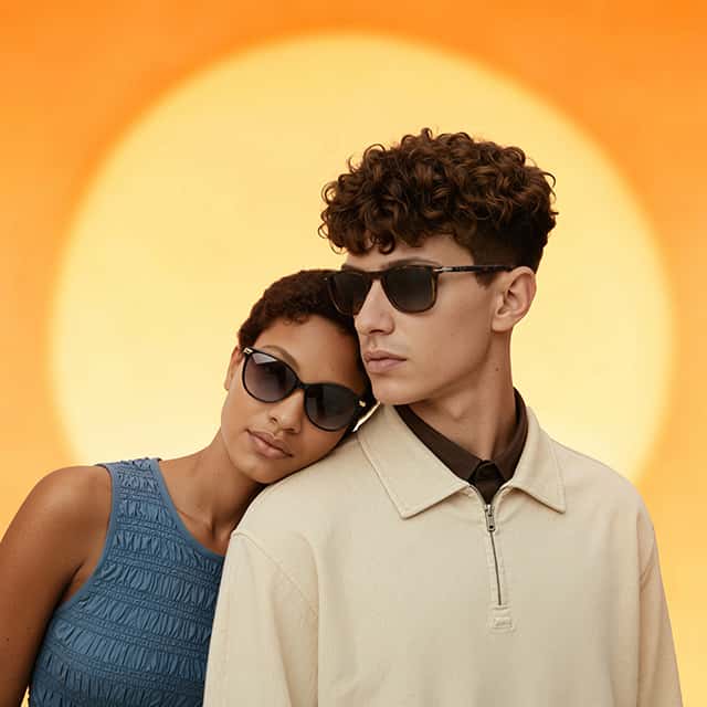 A woman in a blue textured sleeveless top rests her head on the shoulder of a man in a beige jacket. Both are wearing dark sunglasses. They stand against a backdrop of an oversized, blurred orange and yellow sun, suggesting a sunset or sunrise.