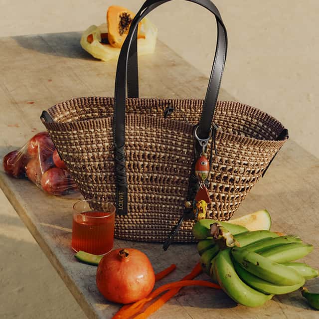 A woven brown handbag with black handles sits on a wooden table. Next to it are small bananas, a whole pomegranate, red onions, and a halved papaya. A glass of orange liquid is placed nearby.