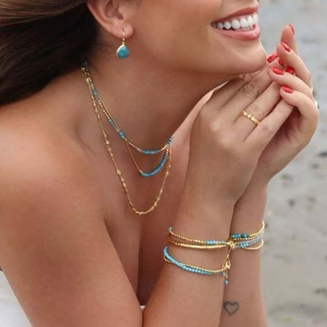 Woman smiling with turquoise jewellery, including necklaces, earrings, and bracelets. She has red nail polish and a small heart tattoo on her wrist, and she's by a beach with sandy shoreline visible in the background.