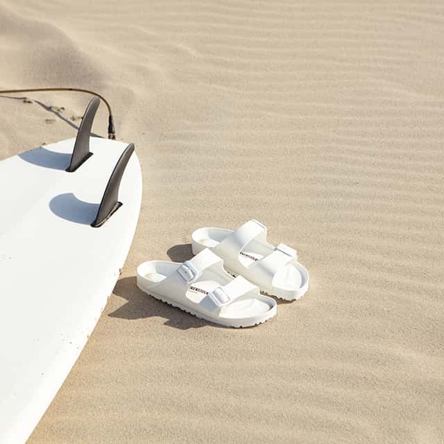 A pair of white sandals rests on smooth sand beside a white surfboard with black fins, suggesting a beach setting.