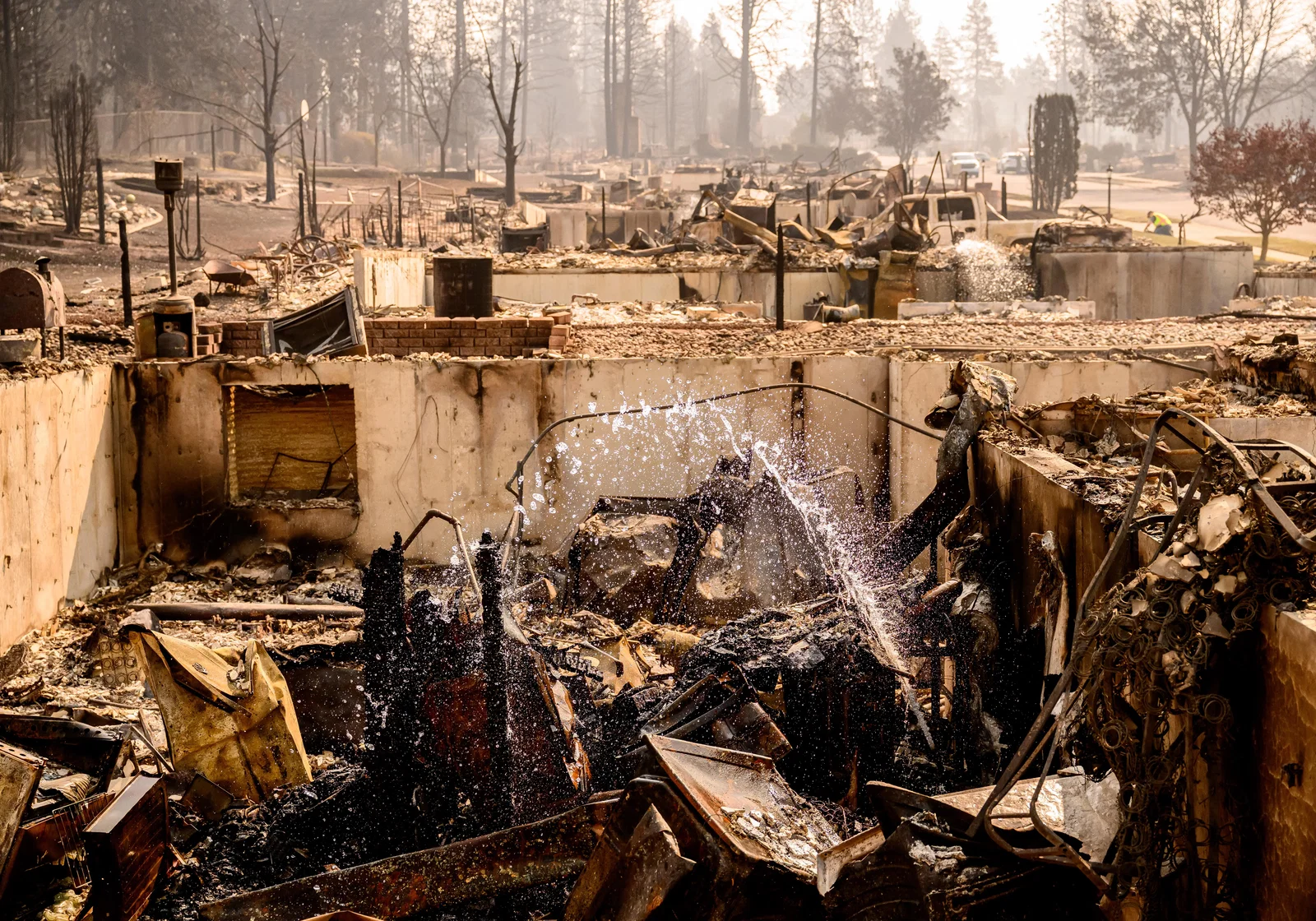 A stream of water arcs over a burnt-down building, with only the foundation remaining under burnt debris.
