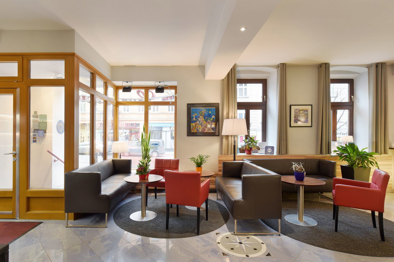 Bright modern hotel lobby with leather sofas, red armchairs, round coffee tables and indoor plants by large street-facing windows