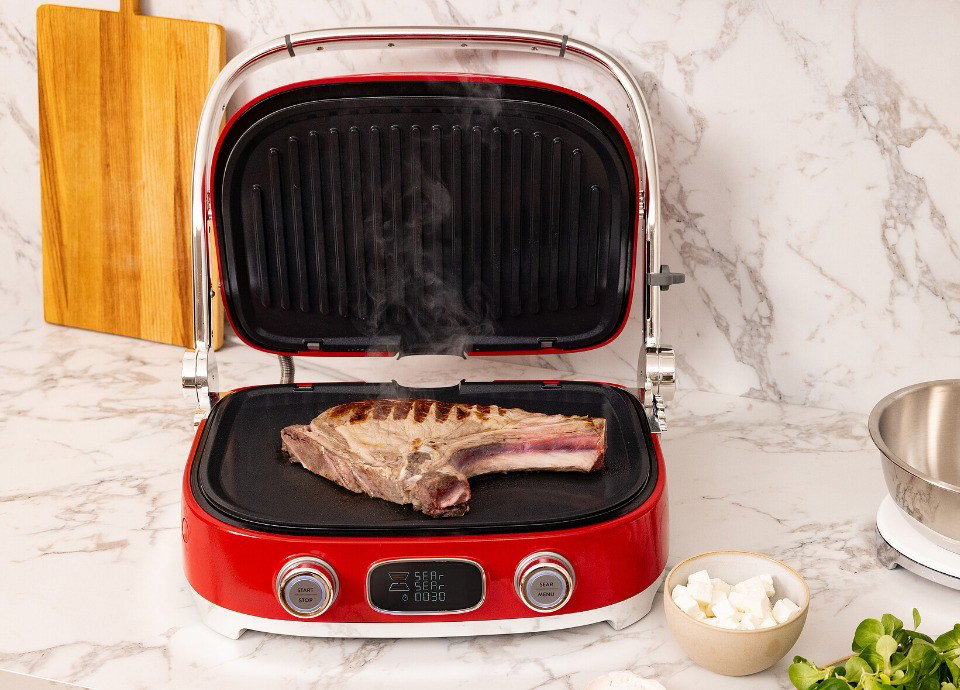 Grill Multiusos Smeg rojo con placas grill cocinando un chuletón sobre una encimera de cocina