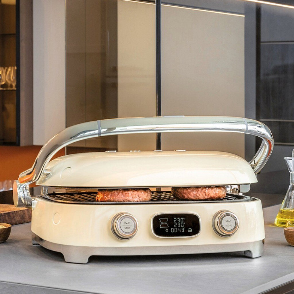 Grill Multiusos Smeg cocinando hamburguesas con la tapa parcialmente cerrada sobre una encimera