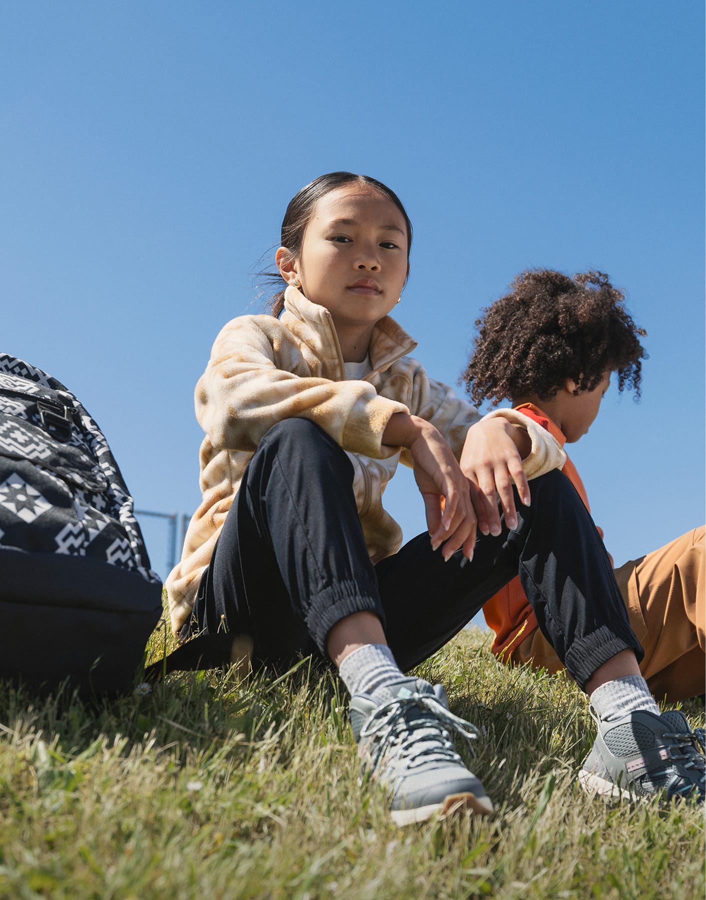 Two kids sitting on a grassy hill with backpacks.