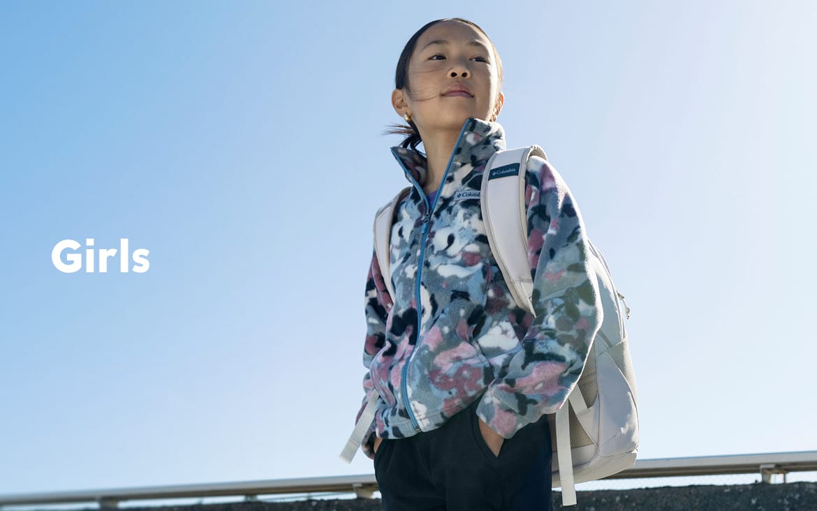 A little girl standing against a blue sky, wearing a backpack with text: Girls