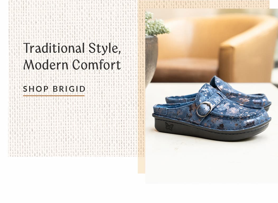 A pair of navy floral Alegria Brigid backless clog slippers with a buckle strap on a light table. Text reads "Traditional Style, Modern Comfort."